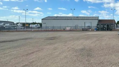 Large gravelled area with grey industrial building and fence in the background