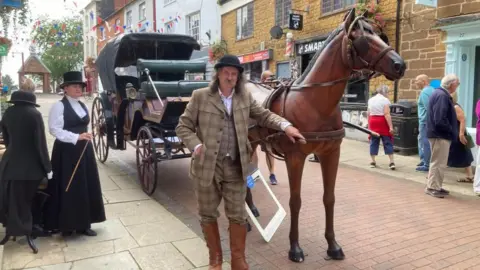 Martin Heath/BBC Sean Lightning in tweed jacket and breeches with a brown bowler hat and brown boots and Alison in top hat and black dress alongside a carriage pulled by a plastic horse