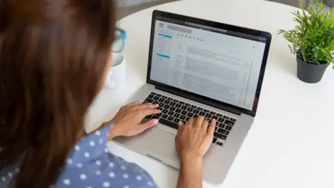 Getty Images Woman in polka-dot blue shirt (with back to camera) reading her e-mails on a laptop. There is a mug on one side of the table and a plant on the other. 