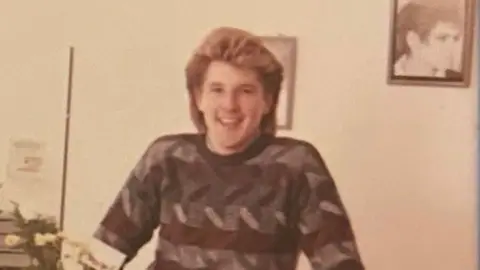 Family photograph Undated image of David Thomas with brown hair with blond highlights wearing a brown and grey patterned jumper standing in one of his hair salons. A framed photograph of a male hair model is on the wall behind him. He is smiling.