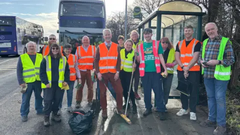 Jacob Bell A group of people in hi-vis jackets stand and smile next to a bus stop. They carry brooms and other cleaning equipment. There are two buses in the background.