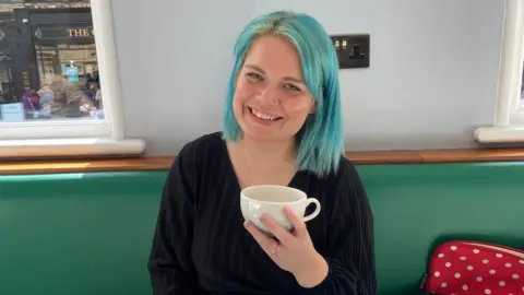 A woman with blue shoulder length hair smiling as she holds up a white mug. She is sitting on a green bench and a shop can be seen behind her. She is wearing a black top.