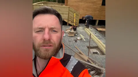 Eddie Riley A man is taking a selfie. He is standing outside and someone is crouching down in the background. He is wearing a bright orange jumper and has dark hair. 