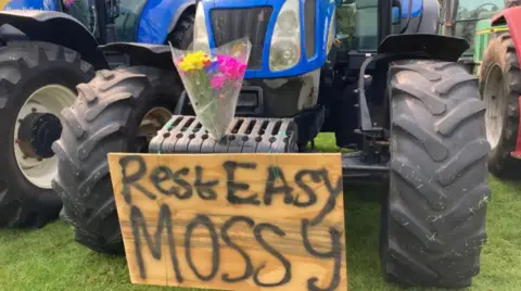 A sign that reads "Rest Easy, Mossy" is on the front of a tractor with a bunch of flowers in tribute to Harry.
