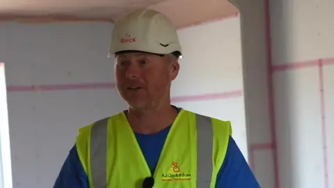 A man in a hard hat and a hi-viz jacket satnds in a partially finished room , the plasterboard is up and the the sheets are bonded by pink scrim tape 