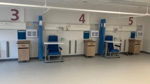 Amy Holmes/BBC A row of numbered hospital treatment bays (3, 4, and 5) lines a clean ward, each equipped with a blue chair, overbed table, medical outlets, and privacy curtains. Small cabinets and wall-mounted equipment sit between the stations under bright ceiling lighting, giving the space an orderly clinical appearance.