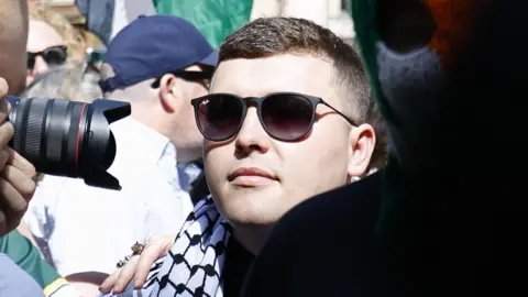 A close-up of a man in the midst of a crowd. He is wearing sunglasses and a black and white keffiyeh headdress around his neck like a scarf. There is a camera lens being held close to his face as a photographer takes a photo