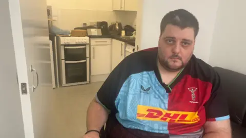 BBC Nathan Rollinson, a man in a red and blue rugby shirt, sitting in front of his kitchen with a cooker that opens onto the floor