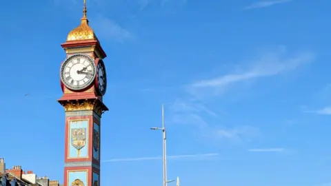 Weymouth Town Council Weymouth's Jubilee Clock on the seafront with the promenade and beach seen behind it. The clock is red and pale blue with gold decoration. The face of the clock is lit with red lights.