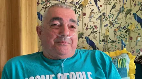 Ally Van Tillo, a grey-haired man with dark eyebrows, looks at the camera while sitting in a lounge chair. He wears a turquoise Stonewall T-shirt