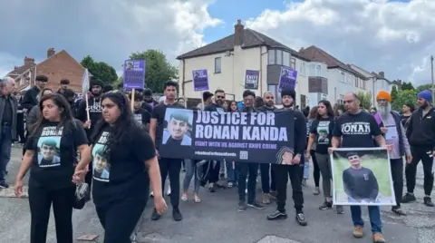 A crowd of people gather for a march. Most people are wearing black t-shirts with a picture of Ronan on the front. A large purple banner with the words Justice for Ronan Kanda is being held up by some of the group.