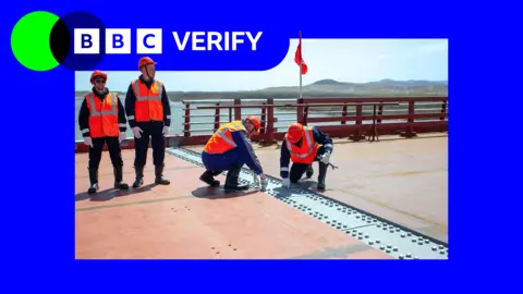 Four workmen wearing bright orange hats and reflective high-vis jackets stand at the centre of a new road bridge between Russia and North Korea. The centrepoint where the two ends of the bridge join is marked by a line of steel plates. Two of the workmen are crouched over the plates, examining them. 