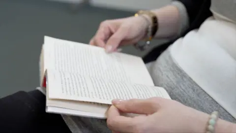 A woman is holding a book in her lap. You cannot see above her stomach. She's holding the book open with both hands and it appears that she is reading the book.
