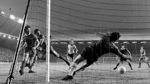 Getty Images A ball shoots just under a diving Parkes at Anfield. Six players are clustered around the goalmouth, with the packed stands in the background