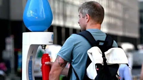 Back view of a man wearing a blue t-shirt and black backpack filling up a red water bottle at a temporary drinking fountain