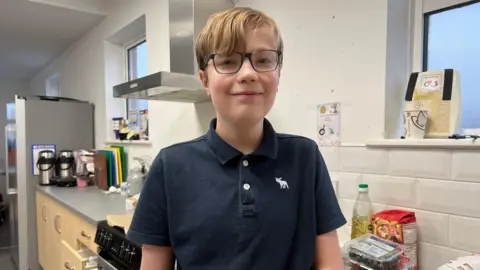 Ellis is wearing a dark blue polo shirt and is stood in a kitchen. He has blonde hair and black glasses