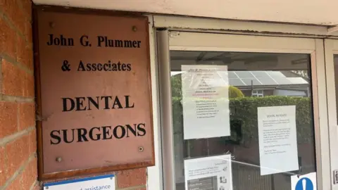 Andrew Turner/BBC A copper-coloured engraved sign reading: "John G Plummer and Associates Dental Surgeons" mounted on a wooden board, outside the premises. Some of the brickwork of the building can be seen in the leftmost side of the image and some signs, explaining the temporary closure and other messages, are stuck to the inside of the glazed panel of an aluminium-framed door.