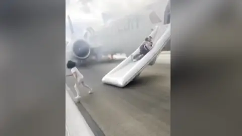 A man slides down an inflatable evacuation slide from a plane as a carrying a child. Another child runs away from the slide. There is smoke in the background, coming from the plane.
