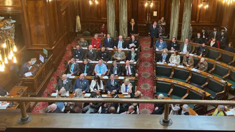 A council chamber with most of the seats full and the focus on a bank of benches with 28 people in them.