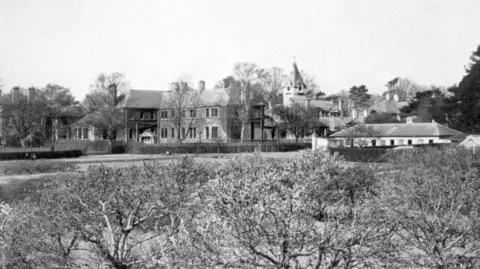 A black and white image of the hospital from the distance. A Victorian looking building can be seen from behind some bushes.