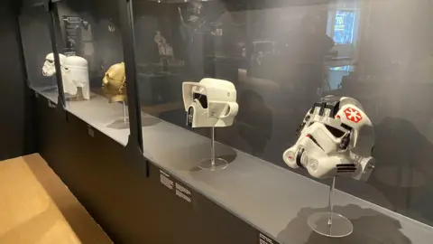 Five helmets in a glass case. They include a white Stormtrooper mask and the gold mask of droid C3PO. There are also three variants of the Stormtrooper helmet as appearing in the films. They have curved bottom halves, and more angular tops around the forehead area. There are eyes in the shape of dark lenses, and in the centre of the helmets, around the mouth area of a wearer, there are grills in the shape of an arrow head, which makes it look like the helmets are frowning.