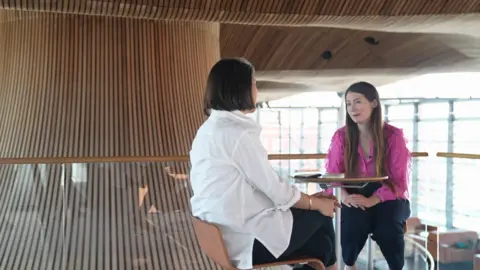 A woman sits at a table facing the camera. She has long, light brown hair and a pink top. She is being interviewed by a reporter in a white shirt who is also sitting at the table, facing her.