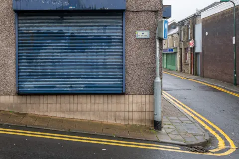A blue shutter down over a shop on a street corner. The building is covered in beige pebble dash and tiles. 
