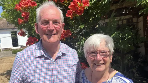 John Devine/BBC Anthony and Helen Chandler are standing in a garden next to a fence which has flowers growing around it. Anthony, who is on the left, has short, white hair and is wearing clear-framed glasses and a blue and red chequered top. Helen, who is on the right, has short, white hair and is wearing pink-framed glasses and a blue and white patterned T-shirt.