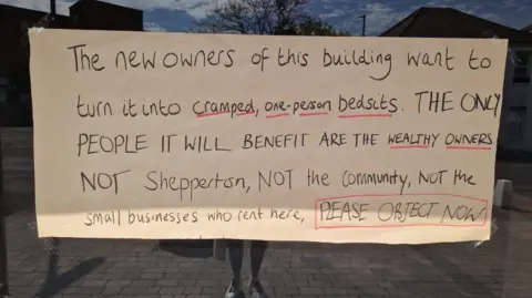 Miranda Schunke/BBC A handwritten sign in a window. It reads "The new owners of this building want to turn it into cramped, one-person bedsits. THE ONLY PEOPLE IT WILL BENEFIR ARE THE WEALTHY OWNERS NOT Shepperton, NOT the community, NOT the small businesses who rent here, PLEASE OBJETC NOW".