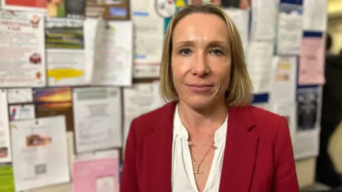 Helen Morgan has a blonde bob and is wearing a white shirt with a red suit blazer. She has a gold necklace on and is standing in front of a board with dozens of posters and leaflets pinned to it.