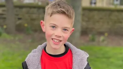 A young blond boy stands on the grass in a park and smiles at the camera. He is wearing a dark red t-shirt and a black coat over it.