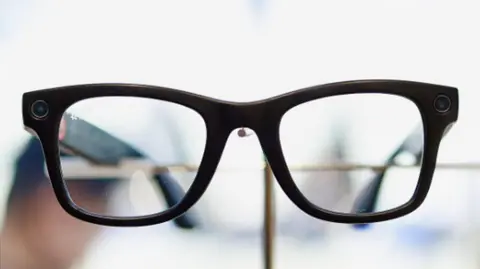A close up of a pair of black smart glasses on a white background . A blurred hand in the baclkground holding the galsses can be seen