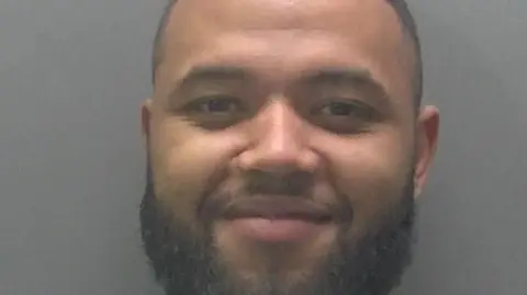 Kane, a man, with black short hair and black beard, smiling for the camera.