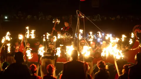Reuters Junior Jarl Squad Vikings with flaming torches surround one boy with a shield who is  standing raised up in the centre. He is carrying a shield and a flaming torch. They are all wearing Viking costumes.