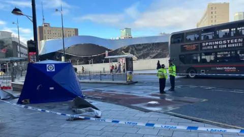 A blue tent on a pavement, surrounded by blue and white police tape. There are two police officers in yellow hi-vis and a black hat standing as scene guards. There is a bus driving past