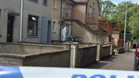 A forensic investigator standing outside a row of terraced properties at Elmfield Walk in Donaghadee where Sarah Montgomery died on Saturday. The investigator is dressed in a white hazmat suit and is holding a clipboard. The properties are two stories in height and there are a couple of sets of concrete steps leading to the first floor properties. There are cordons to the front and side of one ground floor property.