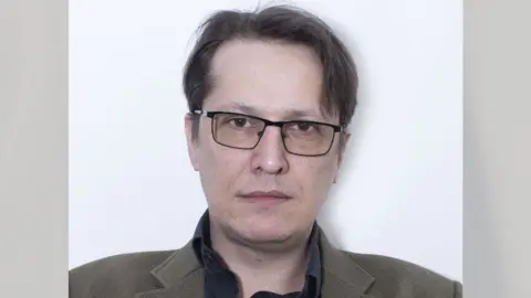 Free Speech Union A head and shoulders shot of Stephen O'Grady looking directly down the camera. He is standing against a white background and is wearing a brown jacket over a dark top. He is clean-shaven and is wearing glasses with black, metal, rectangular frames. His dark brown hair is parted on one side.