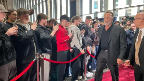 Arnold Schwarzenegger walks on a red carpet in front of a large crowd of students and fans in a glass-fronted university building in Belfast. He has grey hair and a white beard and is wearing a black suit and a black shirt.