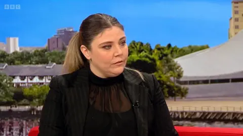 A woman with long brown hair and wearing a black top and black suit jacket is sitting on the red BBC Breakfast sofa. Images of Media City in Salford can be seen behind her.