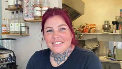 A woman with dyed-pink hair and a blue flower neck tattoo standing next to a silver coffee-making machine.