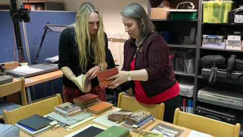 Joanne Parker and Caroline Walter flick through books from their collections which are spread out on a table in piles in front of them. 