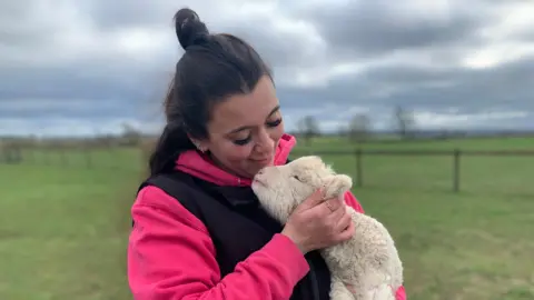 The shepherdess is wearing a pink sweatshirt and a black gilet. She is holding a lamb in her arms and is looking down at it, as the lamb looks up at her.