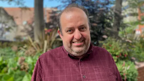 A man with dark hair smiles at the camera. He is wearing a maroon shirt, and behind him are two tress, some bushes, and some red-brick buildings. 