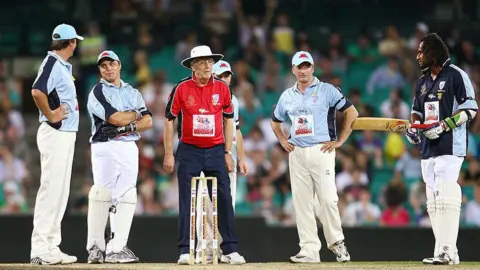 Getty Images  Sir Michael Parkinson - wearing a red shirt and blue trousers and a white hat - stands behind stumps, with five players surrounding him at the Sydney match in February 2009