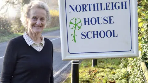 Northleigh House  Viv Morgan outside the school sign looking at the camera and smiling. 