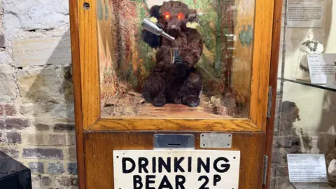 A brown bear toy in a wood and glass case. The bear, which has lit-up red eyes, appears to be pouring contents of a silver bottle into a silver cup. There is a sign under the case that reads 'Drinking Bear 2p'.