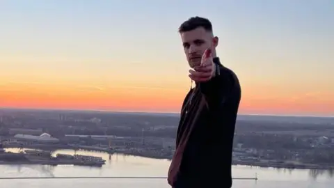 Instagram Lewis Stevenson pointing to the camera on top of a tall chimney in Riga, Latvia