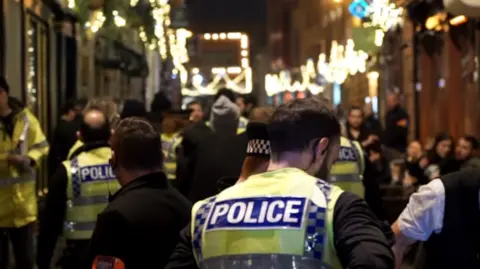 A large number of police officers in high-vis vests are among a crowd of people in a street at night. 