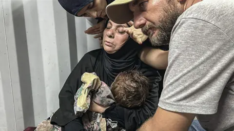 Anadolu caur Getty Images Sieviete, kas valkā melnu hidžābu un abaju, viņas acis aizvērtas, tur bērnu rokās, kamēr vīrietis un sieviete viņu apskāva.