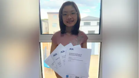 A smiling Ariel with her results. She is holding four sheets of paper. She is in front of a window and buildings are seen behind her. She is wearing glasses.